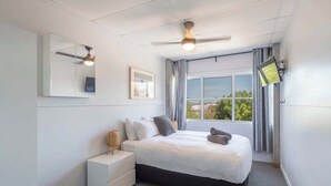 A bedroom with a bed, a nightstand, a ceiling fan, a window with curtains, and a wall-mounted TV.