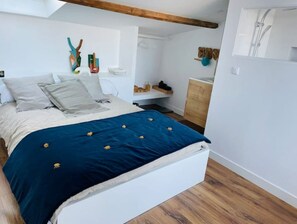 Quarto com cama, piso de madeira, paredes brancas e vigamento de madeira no teto.