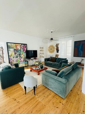 Uma sala de estar moderna com um sofá grande, uma mesa de centro e uma televisão. Há uma estante de livros e um objeto decorativo na parede.