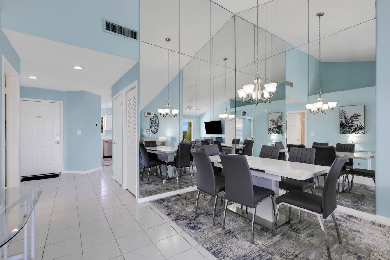 A modern dining area with a large table and chairs, a chandelier, and a large mirror reflecting the room.