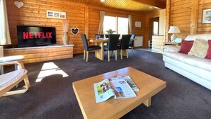 Interior acolhedor de uma cabana de madeira, com teto de madeira, uma televisão identificada como 'NETFLIX', uma área de refeições com cadeiras e uma mesa, e um sofá com almofadas.