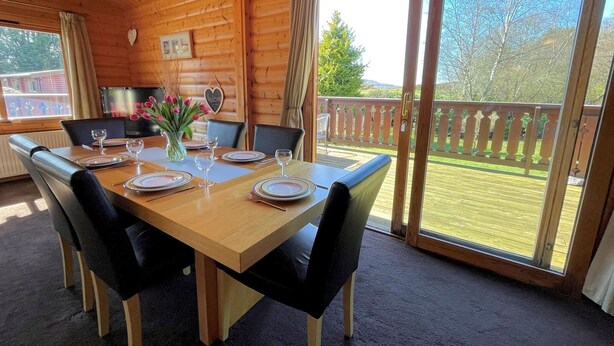 Uma cabana de madeira com mesa de jantar posta para quatro, cadeiras, um vaso de flores e vista para um deck e árvores.