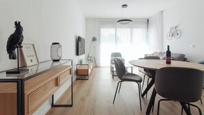 Ein modernes Wohnzimmer mit Essbereich, ausgestattet mit einem Holztisch, schwarzen Stühlen, einer Couch und einem an der Wand montierten Fernseher.
