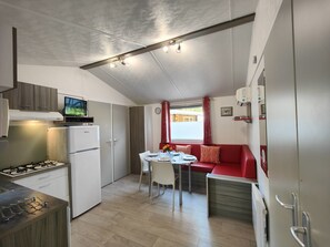 A compact kitchen with a gas stove, a refrigerator, and a television on a cabinet. There is a dining area with a table and chairs, and a red sofa.