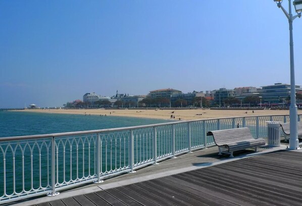 A wooden boardwalk with a metal railing, benches, and a view of a beach and buildings in the background.