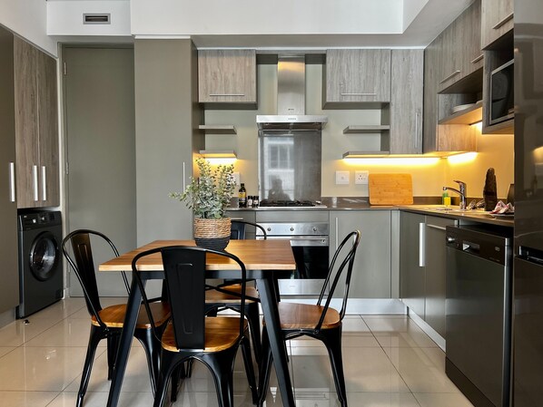 A modern kitchen with built-in appliances, a dining area with a wooden table and chairs, and a potted plant.