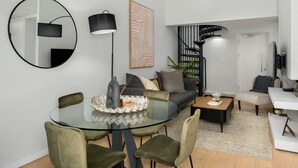 A modern living room with a glass dining table, green upholstered chairs, a black floor lamp, a grey sofa, a coffee table, and a spiral staircase. - Designer Apartment in Sea Point (Cape Town)