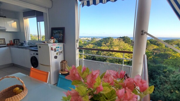 Una habitación con una mesa de comedor, sillas, una cesta y una planta en maceta. Se ve una ventana con vistas a zonas verdes y un edificio a lo lejos.