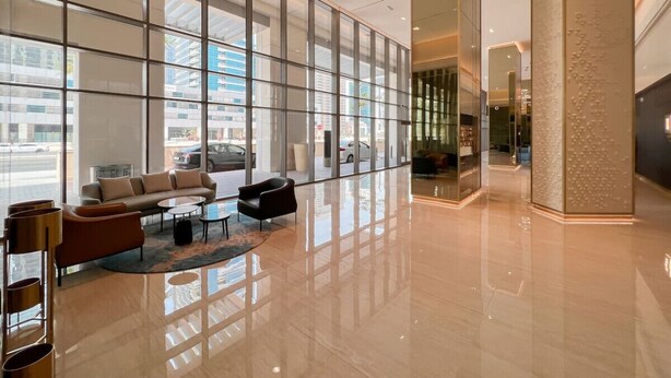 A spacious lobby with a modern design, featuring a seating area with sofas and a coffee table, large glass windows offering a view of the city, and a polished floor reflecting the interior.