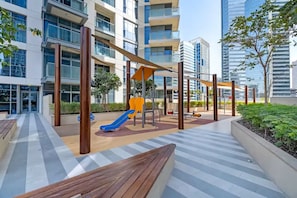 Un espacio exterior moderno con un parque infantil, bancos y un edificio con amplios ventanales.