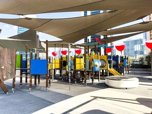 An outdoor playground with various play structures, including slides and climbing frames, under a shaded canopy.