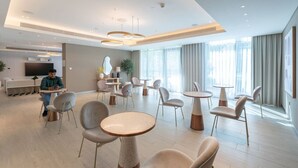 Eine moderne Hotelhalle mit runden Tischen und Stühlen, einem Flachbildfernseher und großen Fenstern mit Vorhängen.