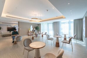 Eine moderne Hotelhalle mit runden Tischen und Stühlen, einem Flachbildfernseher und großen Fenstern mit Vorhängen.