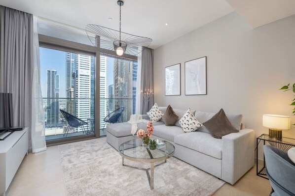 A modern living room with a grey sofa, a glass coffee table, and a city view through large windows.