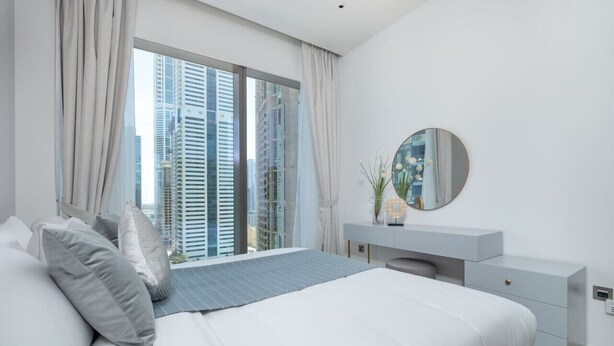 A modern bedroom with a large bed, a vanity with a mirror, and a view of tall buildings through large windows. - Vogue, Marina Gate 2 near JBR Beach and Marina Mall (Dubai)