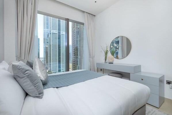 Ein modernes Schlafzimmer mit einem großen Bett, einem Schminktisch mit Spiegel und Blick auf hohe Gebäude durch große Fenster.