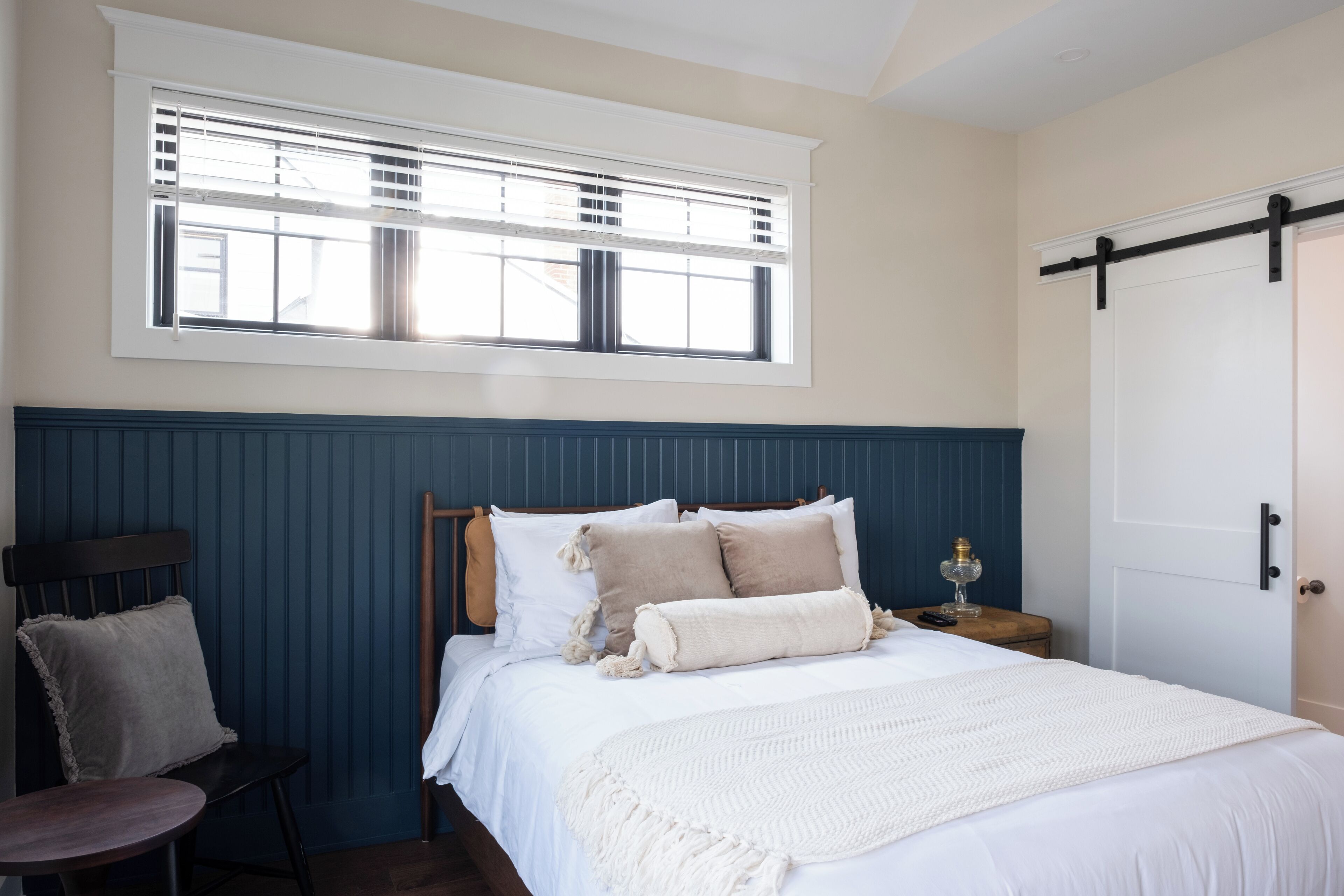 A bedroom with a bed, a chair, a bedside table, a lamp, and a window with blinds.