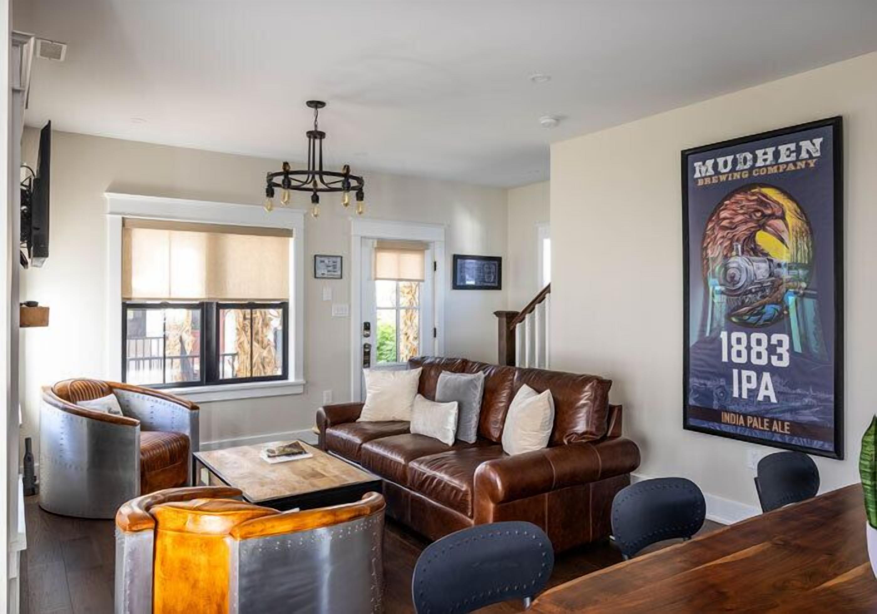 A modern living room with a brown leather sofa, a wooden coffee table, and a chandelier. There is a poster on the wall and a staircase leading to another room.
