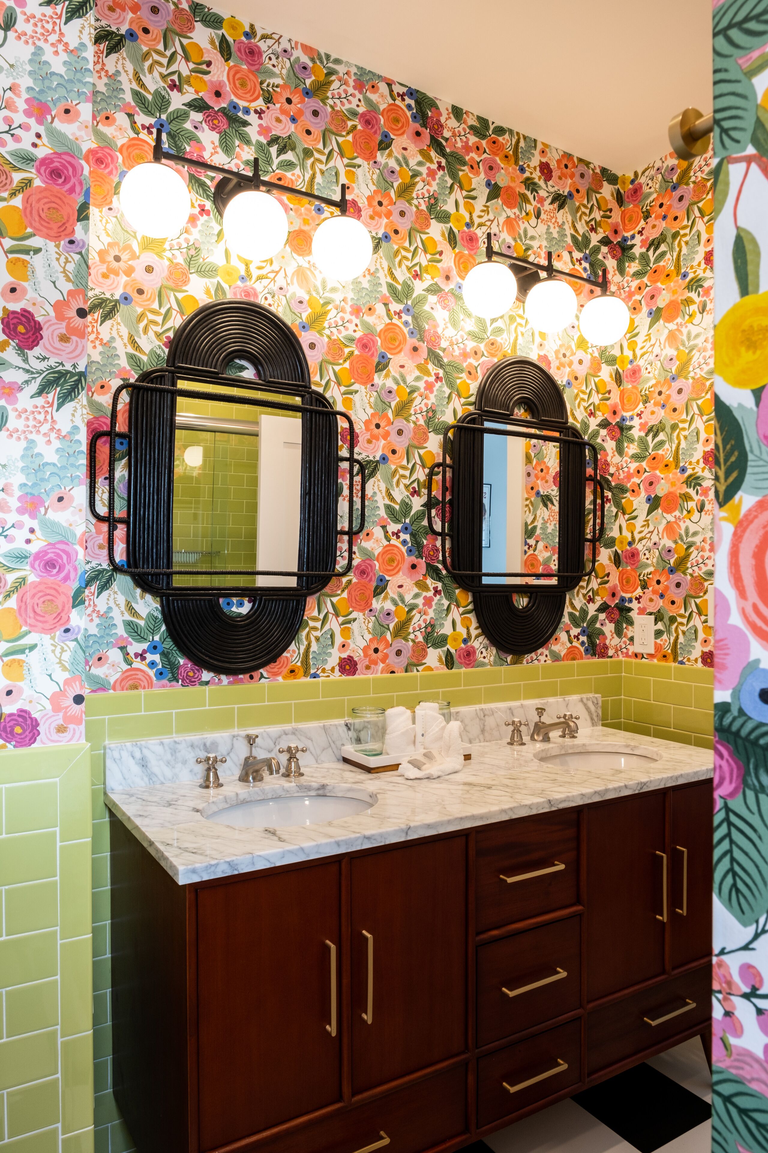 A bathroom with a floral wallpaper, two mirrors with black frames, a marble sink, and wooden cabinets with gold handles.