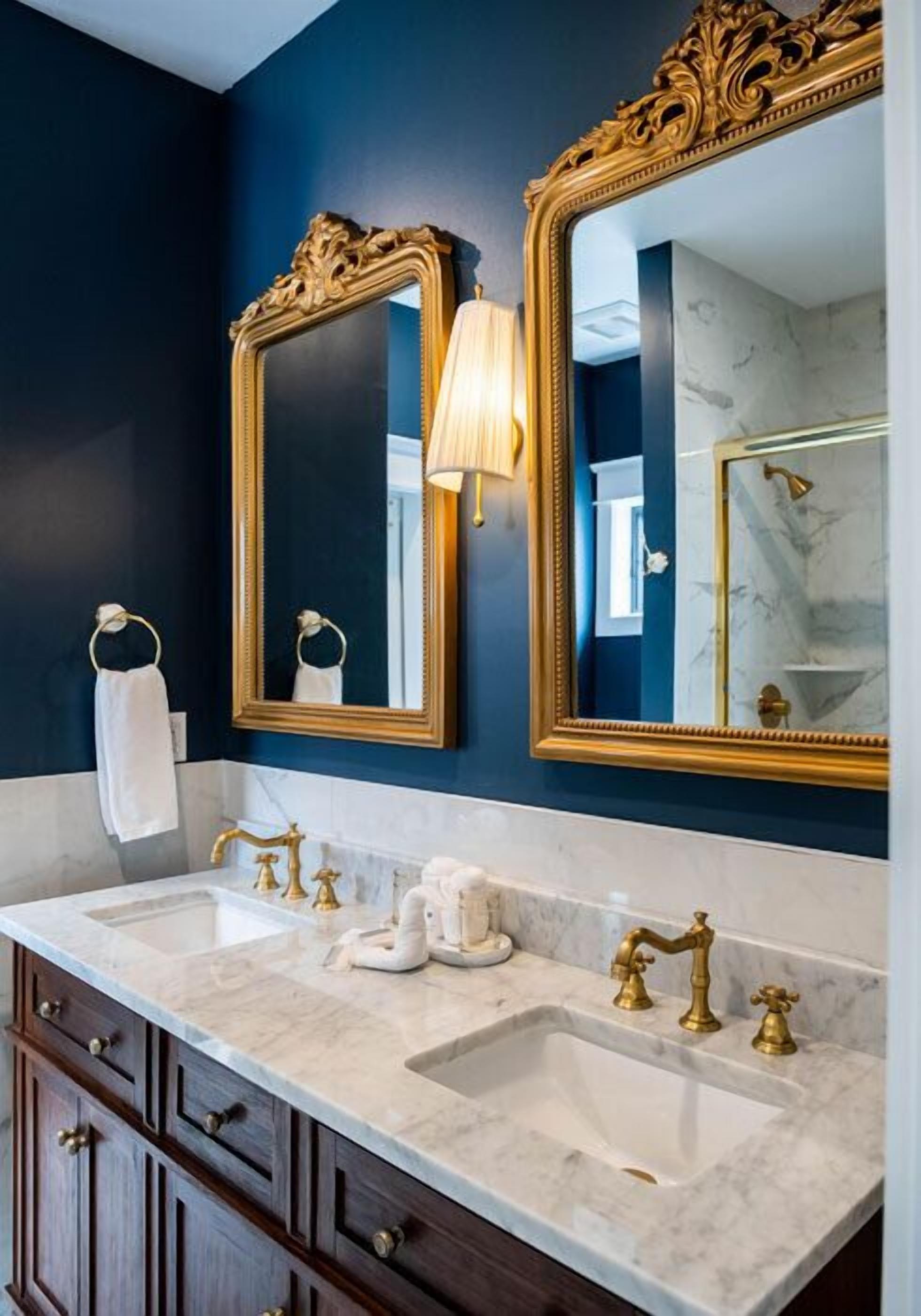 A bathroom with dark blue walls, two gold-framed mirrors, a white marble sink, and a dark wooden cabinet.