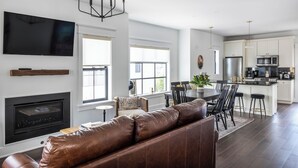 A modern living room with a leather sofa, a fireplace, a dining area with a table and chairs, and a kitchen with white cabinets and appliances. - The Hen Houses (Wildwood)