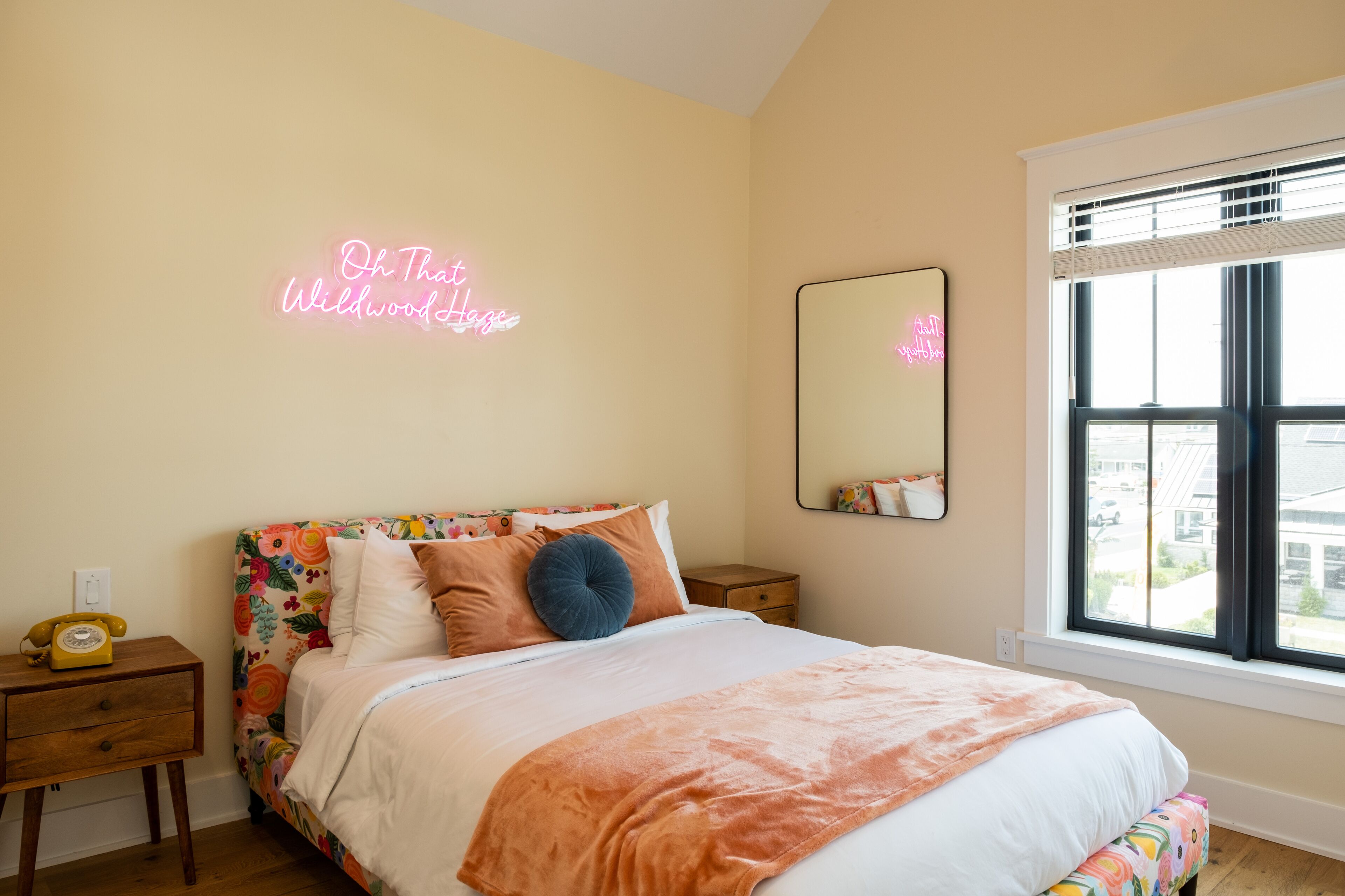 A bedroom with a floral headboard, a neon sign, a bedside mirror, and a window with a view.