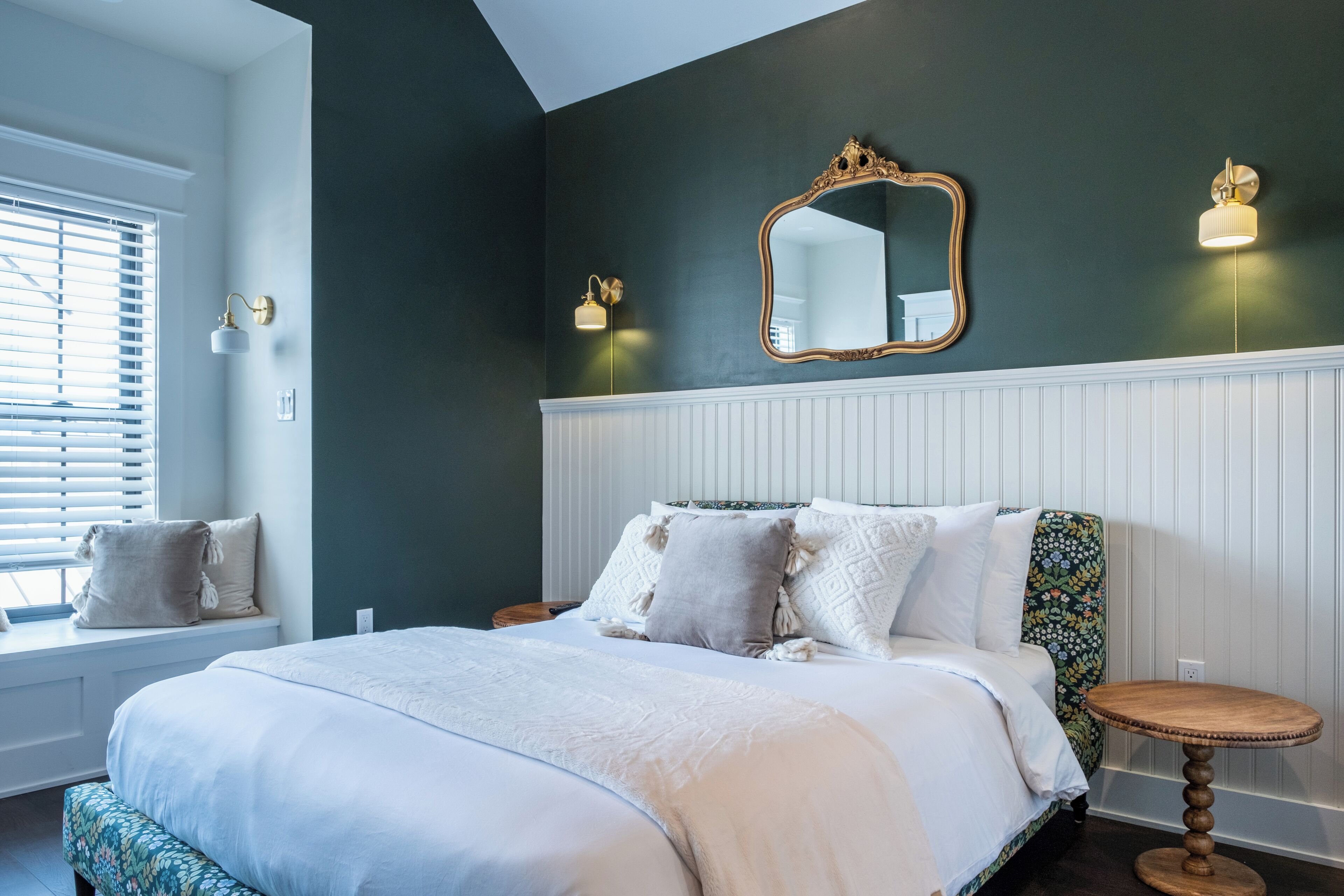 A bedroom with a large bed, a window with blinds, a round wooden side table, and a mirror on the wall.