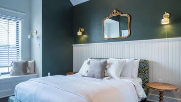 A bedroom with a large bed, a window with blinds, a round wooden side table, and a mirror on the wall.