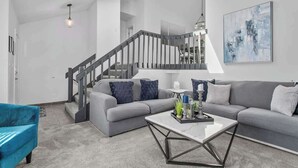 A modern living room with a grey sofa, blue armchair, and a coffee table. A staircase with a grey railing is visible in the background.