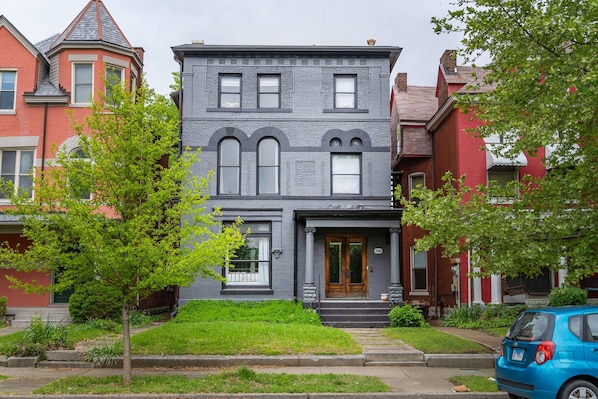 A two-story gray townhouse with a front porch, a small front yard, and a blue car parked on the street.