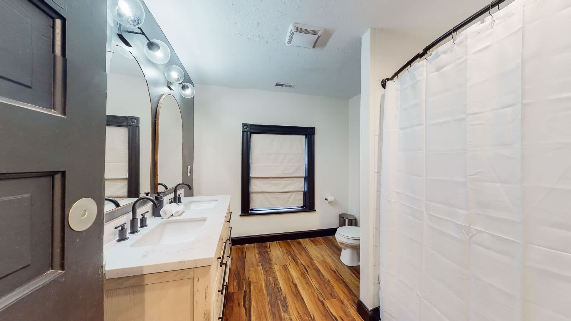 A bathroom with a wooden floor, a white sink, a mirror with lights, a window with a towel, and a shower curtain.