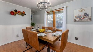Un comedor con una mesa de madera dispuesta para cuatro personas, rodeada de sillas marrones. Un candelabro cuelga del techo, y se ve una ventana con cortinas blancas.