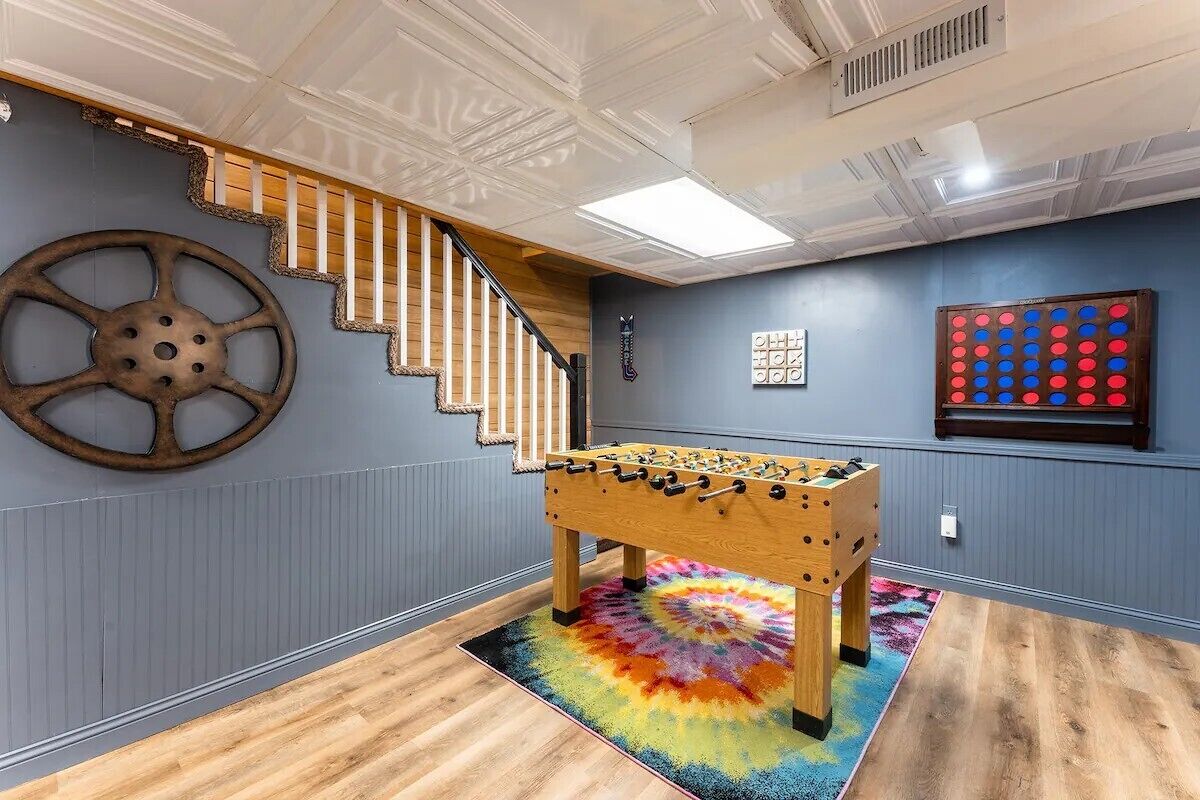 Una habitación con una mesa de futbolín de madera, un tapete de estampado jaspeado y lleno de color, una decoración de rueda grande de madera, una escalera y un tablero de juego montado en la pared.