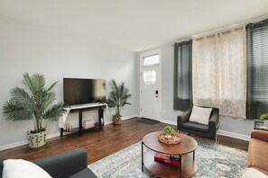A living room with a flat-screen TV, two potted plants, a coffee table with a book, a sofa, and a window with curtains.