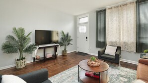 Ein Wohnzimmer mit Flachbildfernseher, zwei Blumentöpfen, einem Couchtisch mit einem Buch, einer Couch und einem Fenster mit Vorhängen.