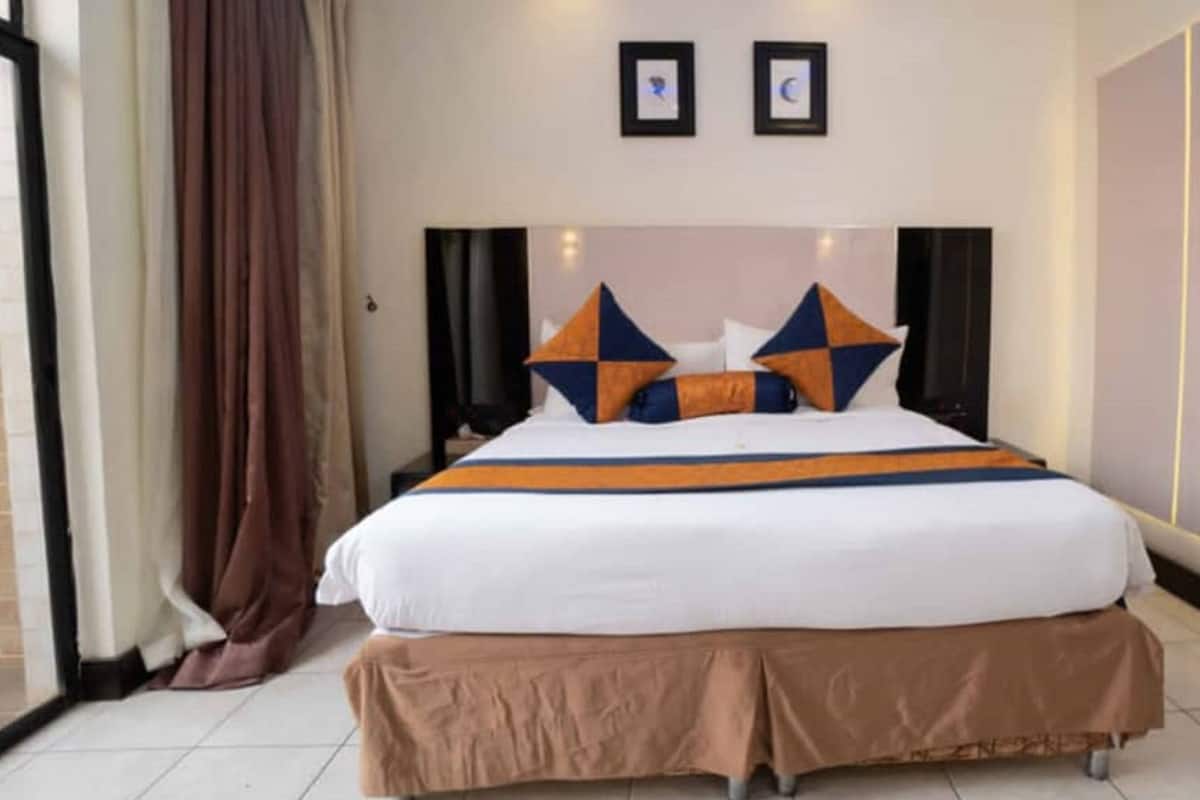 A hotel room with a large bed, a brown skirted bed base, two wall-mounted speakers, and a curtain on the left.