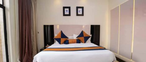 A hotel room with a large bed, a headboard, two wall-mounted picture frames, and a curtain on the left.