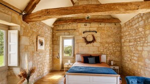 Un dormitorio con paredes de piedra, techo de madera, una cama, una mesita de noche, una silla y un jarrón con flores secas.