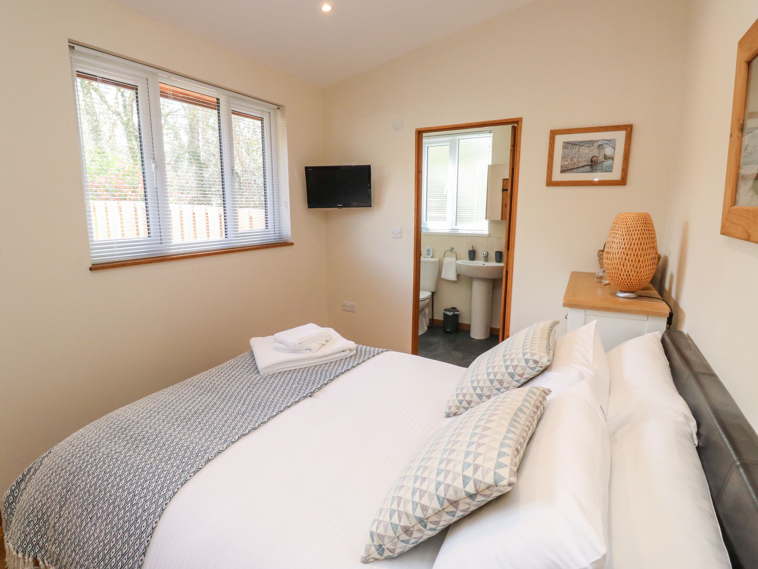 A bedroom with a bed, a TV mounted on the wall, a window with blinds, a bathroom visible through an open door, and a framed picture on the wall.