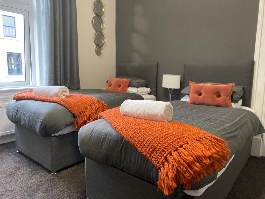 Habitación de hotel con dos camas, ropa de cama gris, mantas naranjas y cabecero gris.