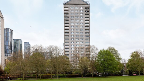 Un grand immeuble d’habitation doté de nombreux balcons, entouré d’un parc verdoyant avec des arbres et une aire de jeux.