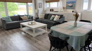A modern living room with a grey sofa, a white coffee table, and a dining table with chairs. There is a kitchen area with cabinets and a window with a view of the outdoors. - All Season Cottage steps to Long Beach (Wainfleet)