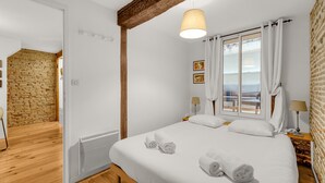 A bedroom with a large bed, white linens, a wooden ceiling beam, a window with curtains, and a view of the sea. - Hippolyte - Appt avec terrasse couverte (Toulouse)