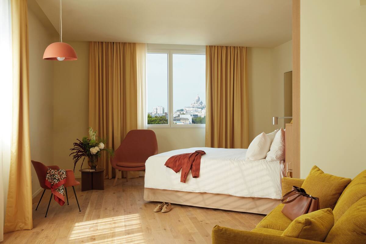 A hotel room with a bed, a chair, a sofa, a window with a city view, and a hanging light fixture.