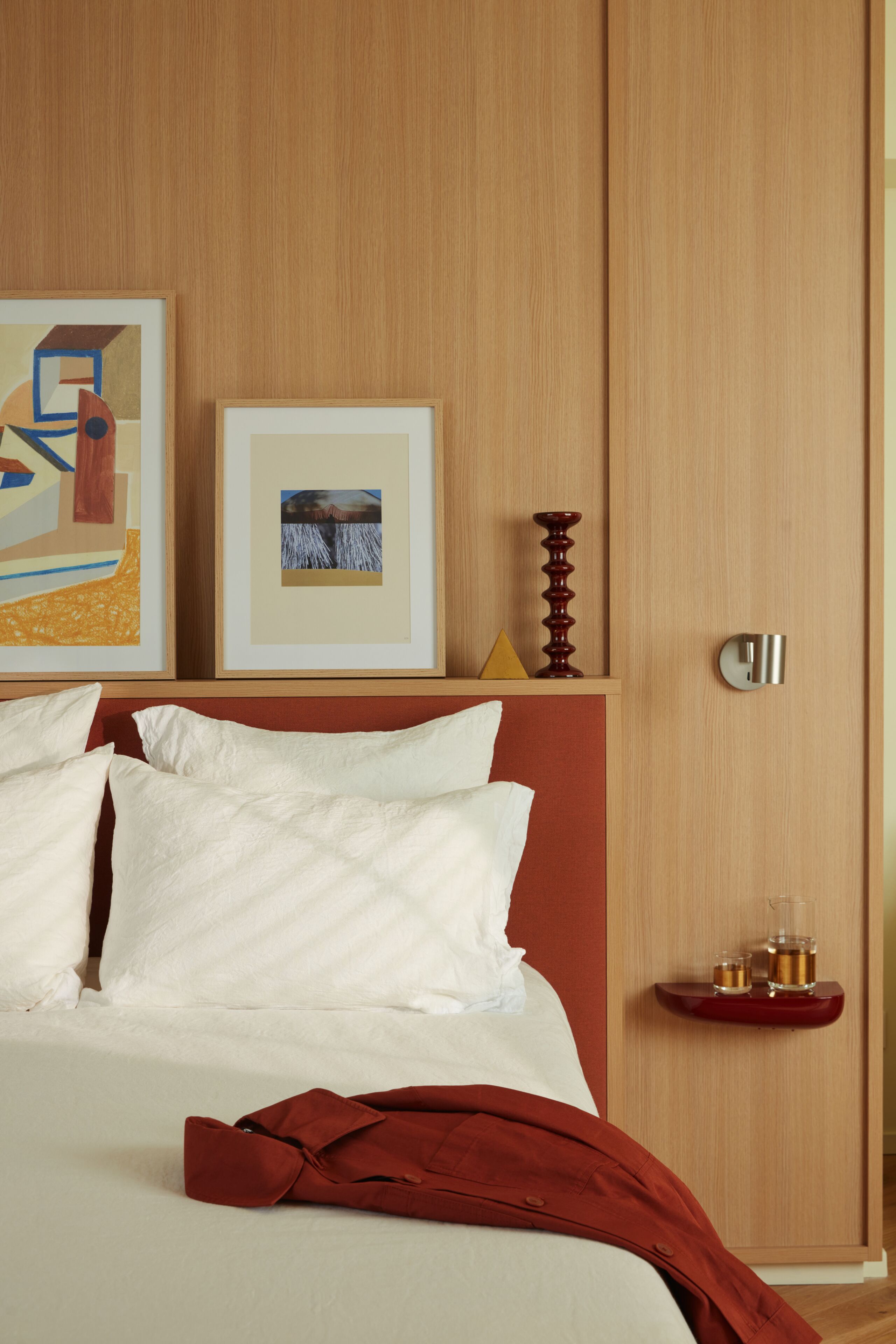 A neatly made bed with white linens and a red jacket draped over it, a wooden headboard, and two framed pictures on the wall.