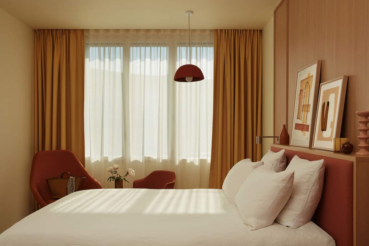 A hotel room with a large bed, a red chair, a small table with a vase of flowers, and a window with curtains.