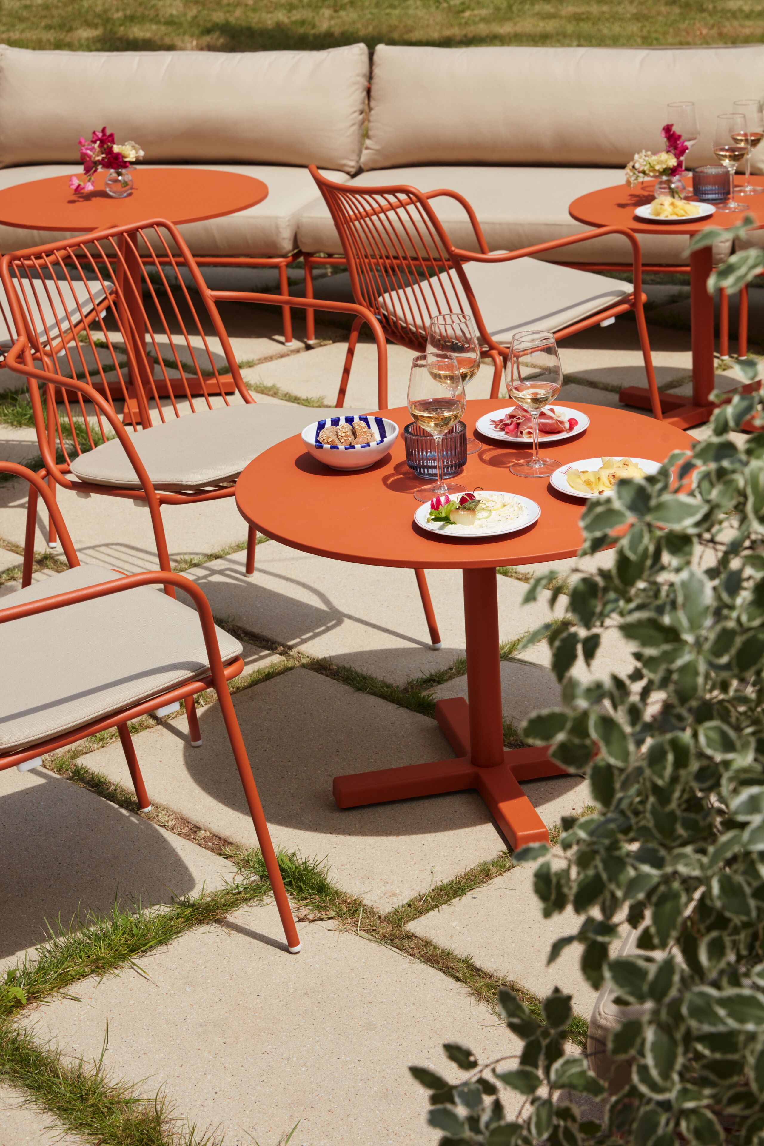 An outdoor dining area with orange metal chairs and tables, a beige sofa, and a small garden.
