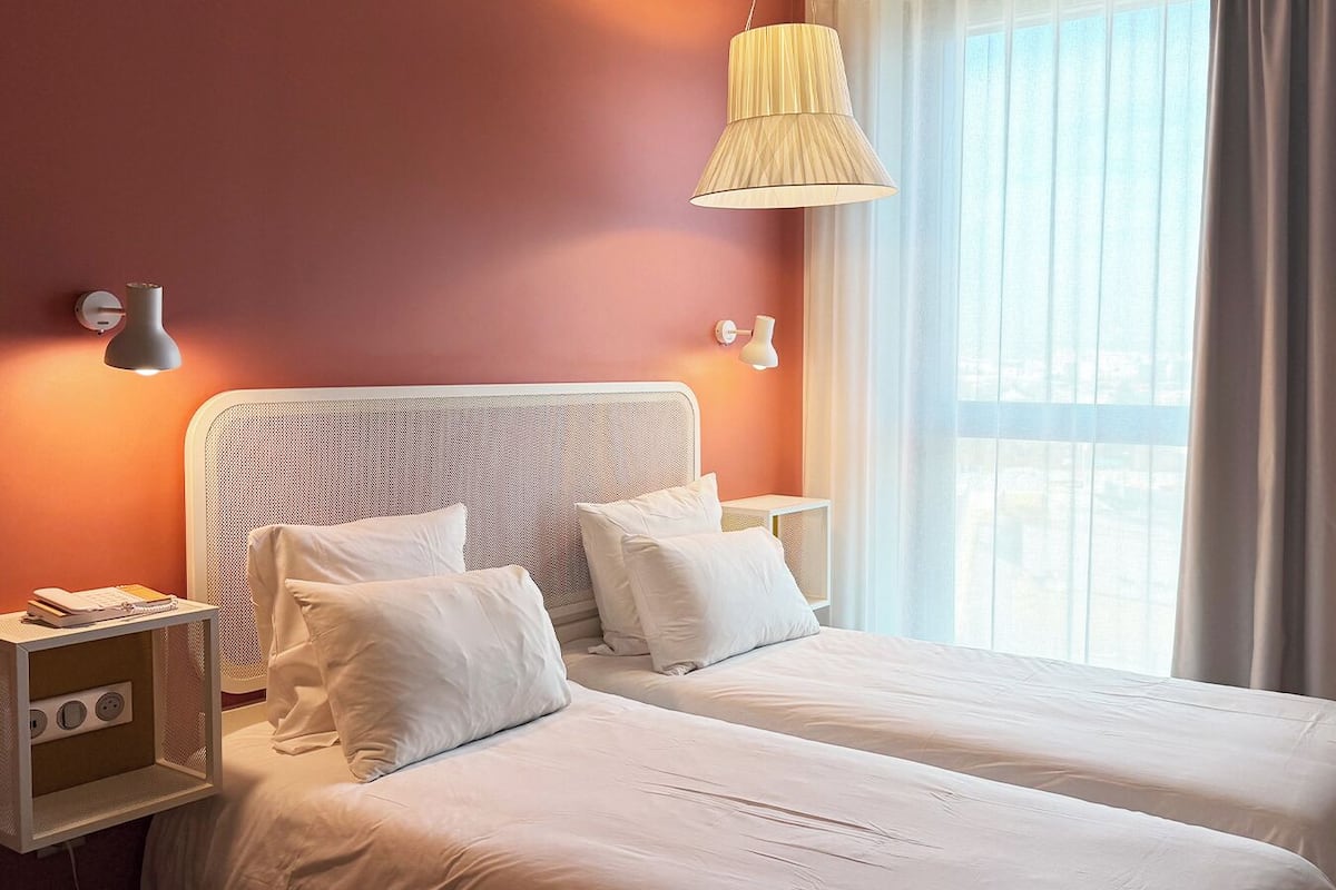 A hotel room with two beds, a large window with curtains, and a hanging lamp.