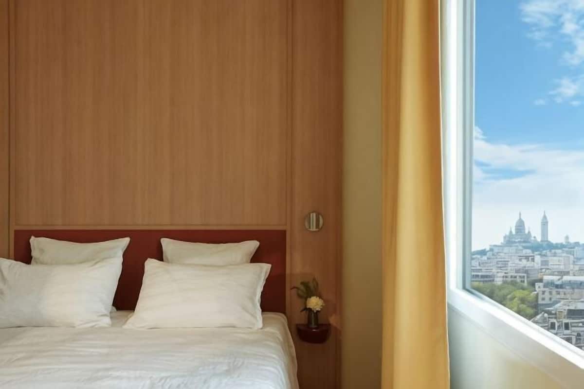 A hotel room with a large bed, a wooden headboard, a nightstand with a vase, and a view of a cityscape through a window.
