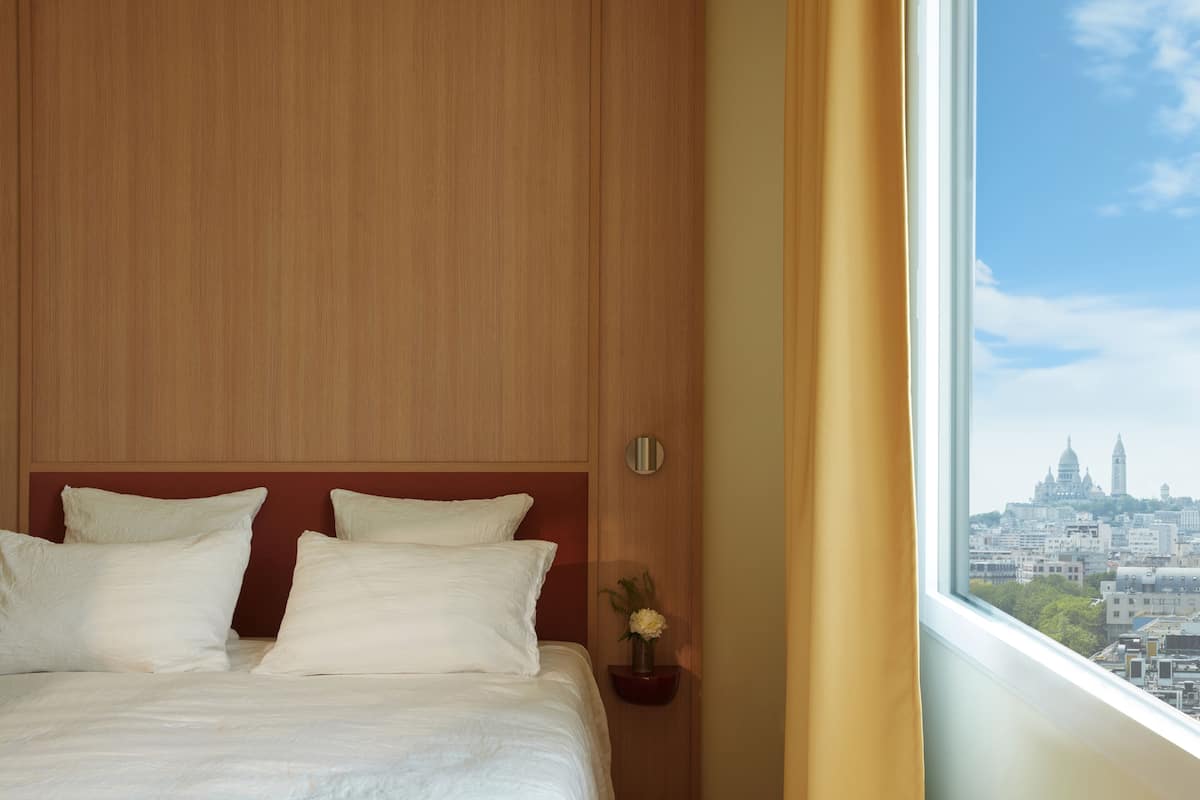 A hotel room with a large bed, wooden headboard, and a view of a cityscape through a window.